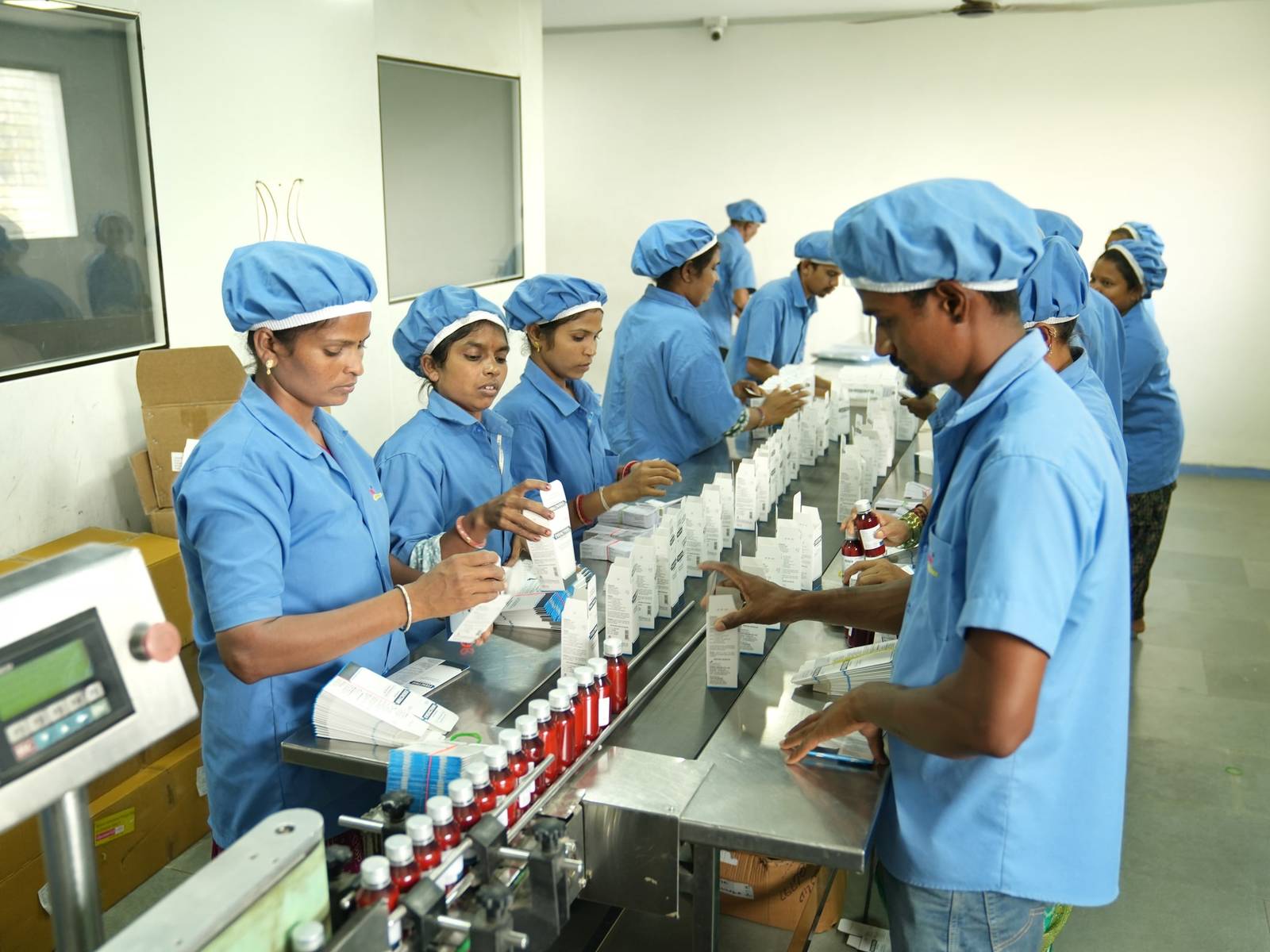 Production staff in blue uniforms and hair caps packing liquid oral bottles into cartons along a stainless-steel packing line